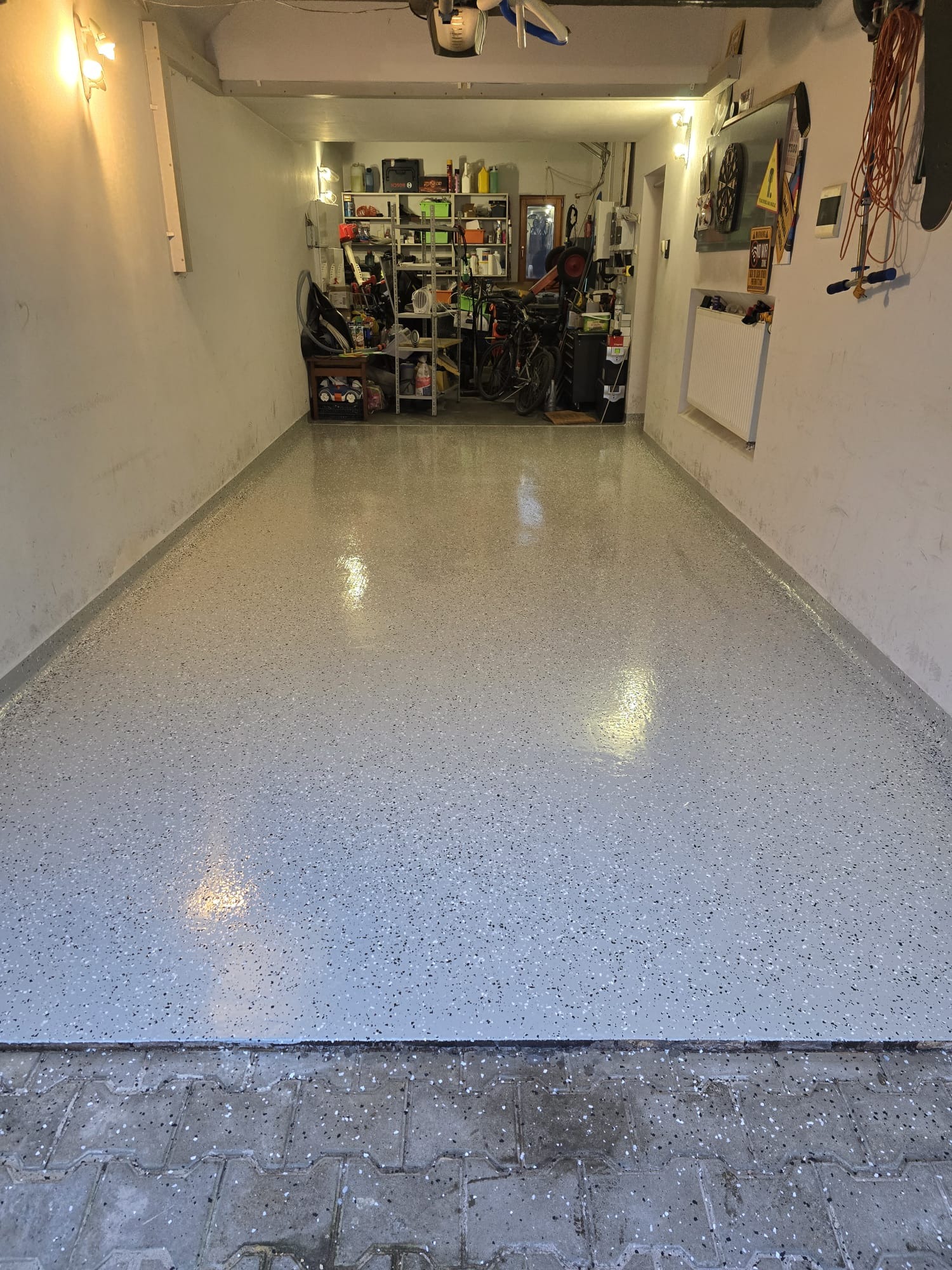 Jasnoszara podłoga w garażu z drobnymi, ciemnymi plamkami, widok z perspektywy wejścia, w tle regały z narzędziami i rower.