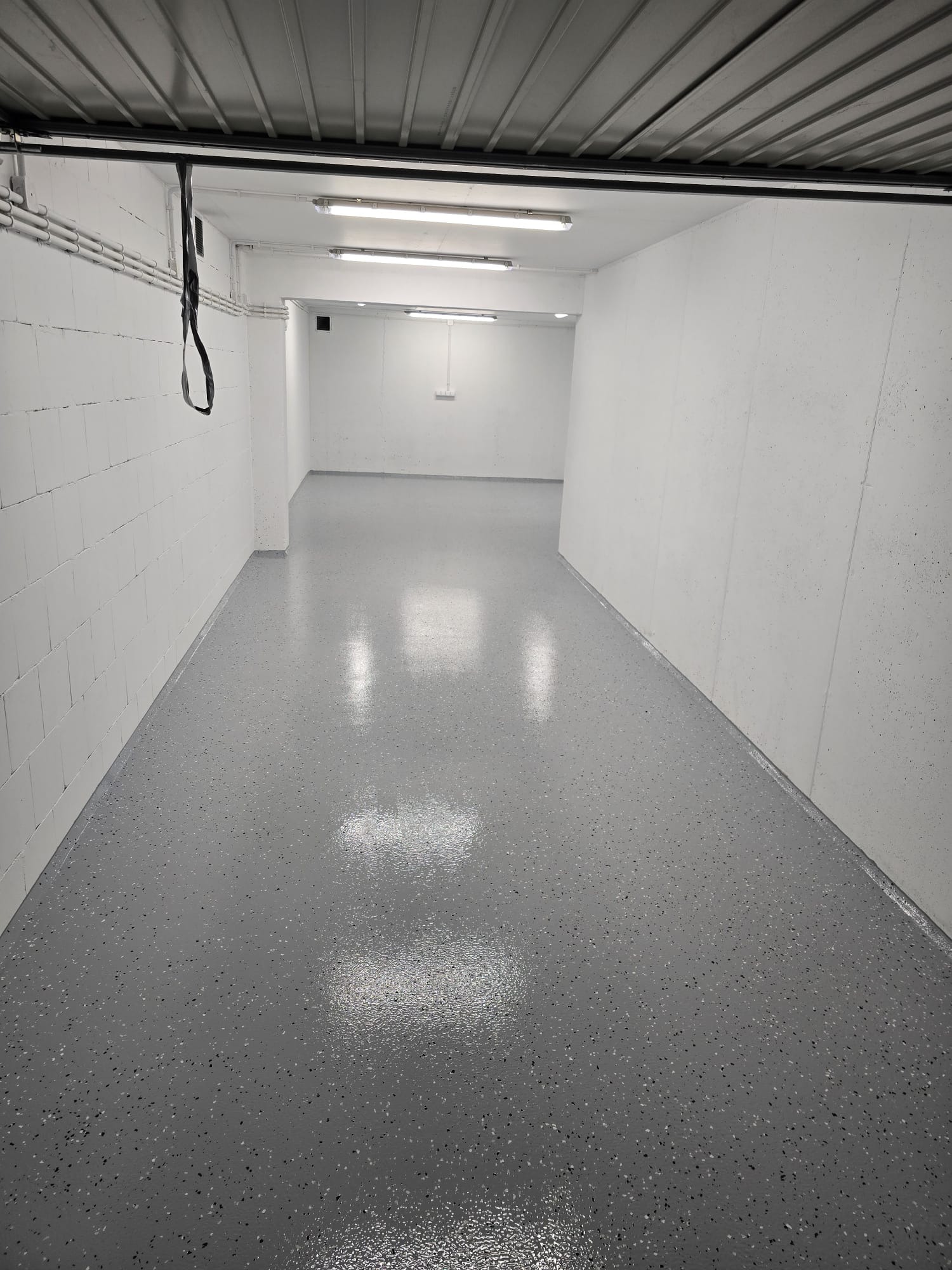 Szara, błyszcząca podłoga w garażu z widocznymi, drobnymi, ciemnymi plamkami, oświetlona przez podłużne lampy sufitowe, z białymi ścianami z betonu komórkowego.
