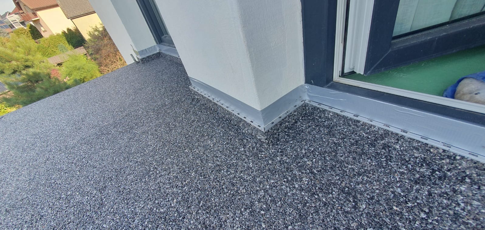 Szara, kamyczkowa nawierzchnia balkonu z widocznymi profilami wykończeniowymi przy ścianie i oknie.
