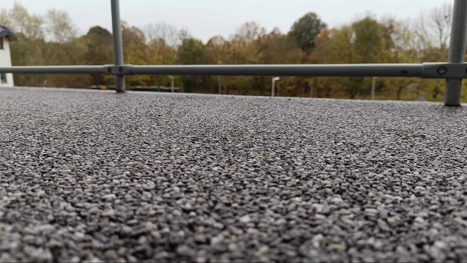 Szara, drobnoziarnista powierzchnia z widocznym metalowym zabezpieczeniem krawędzi i drzewami w tle, sfotografowana z niskiej perspektywy.