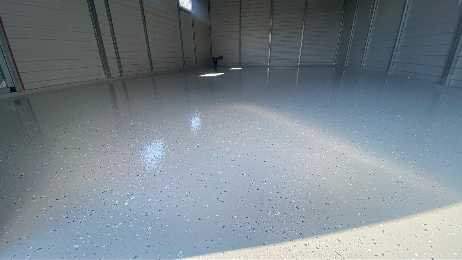 Gładka, jasnoszara podłoga z drobnymi, ciemnymi i jasnymi plamkami wewnątrz metalowego garażu, z widocznym odbiciem światła słonecznego.