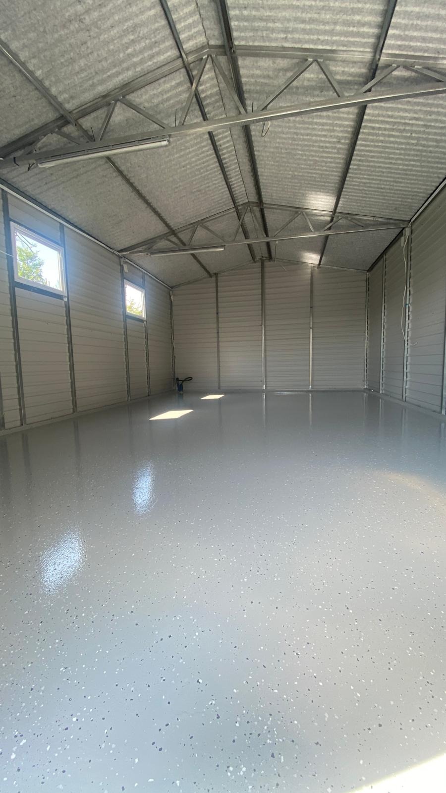 Wnętrze metalowego garażu z nową, jednolitą, jasnoszarą podłogą z drobnymi, ciemniejszymi plamkami, widoczne okna i metalowa konstrukcja dachu.