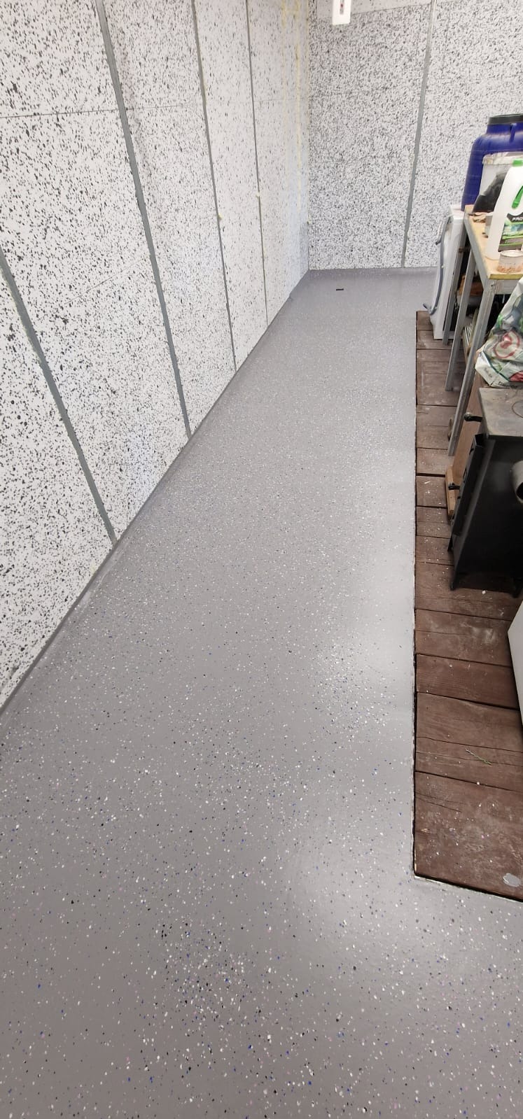 Szara, gładka posadzka z drobnymi, kolorowymi wtrąceniami, przylegająca do ściany z płyt o chropowatej fakturze, częściowo zakryta przez drewniany podest i sprzęty warsztatowe.