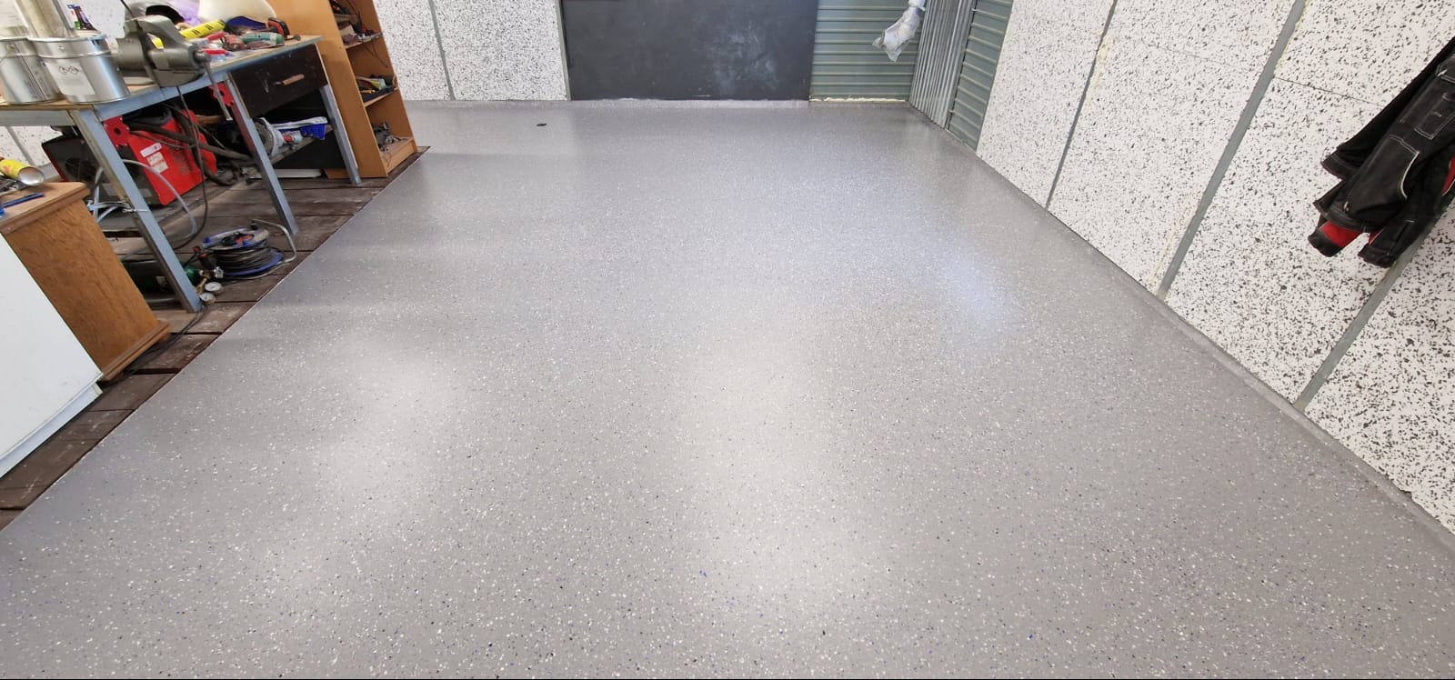 Świeżo wylana, szara posadzka z drobnymi, kolorowymi wtrąceniami w przestronnym pomieszczeniu warsztatowym, widoczne narzędzia i sprzęt w tle.