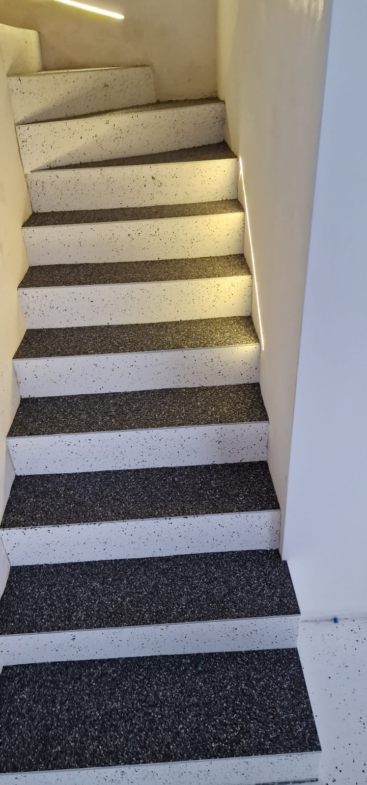 Schody wykończone szarą, nakrapianą posadzką, z subtelnym oświetleniem LED wzdłuż ściany.