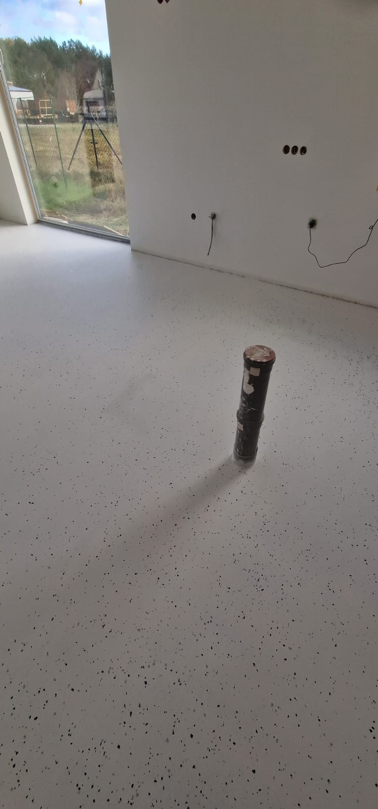 Jasna podłoga z drobnymi, ciemnymi plamkami w nowym domu, widoczne niedokończone gniazdka elektryczne na ścianie i rura wystająca z podłogi.