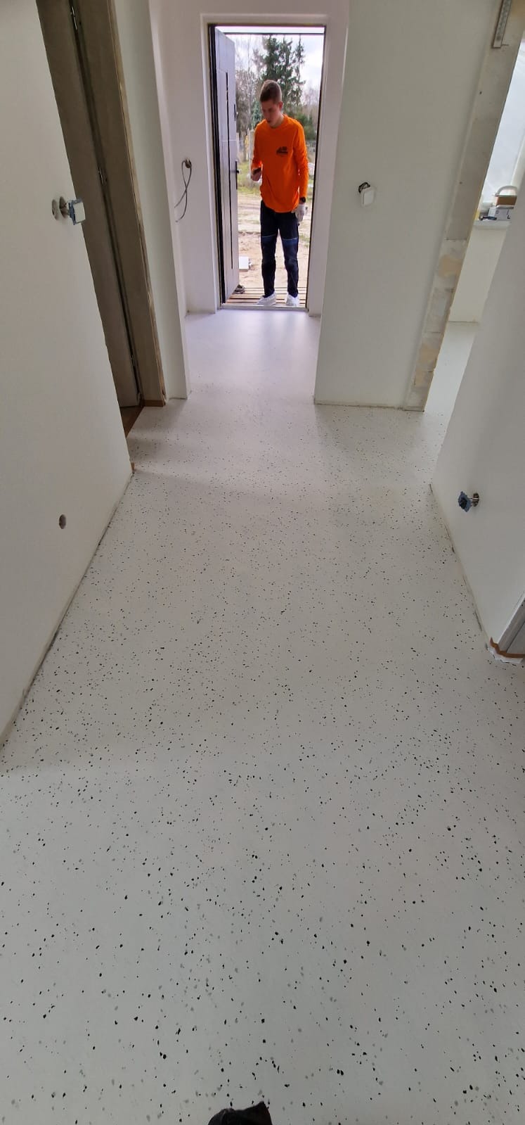 Biała posadzka żywiczna z czarnymi drobnymi plamkami w nowym domu, widoczny pracownik w pomarańczowej bluzie stojący w drzwiach.