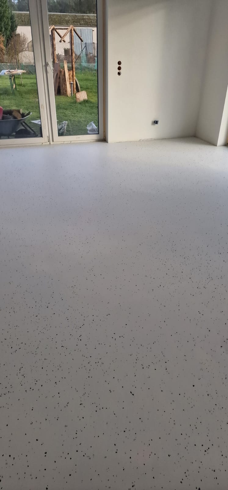 Jasnoszara podłoga z drobnymi czarnymi plamkami, widok na okno z ogrodem i drewnianą konstrukcją w tle, świeżo pomalowane białe ściany z widocznymi gniazdkami elektrycznymi.