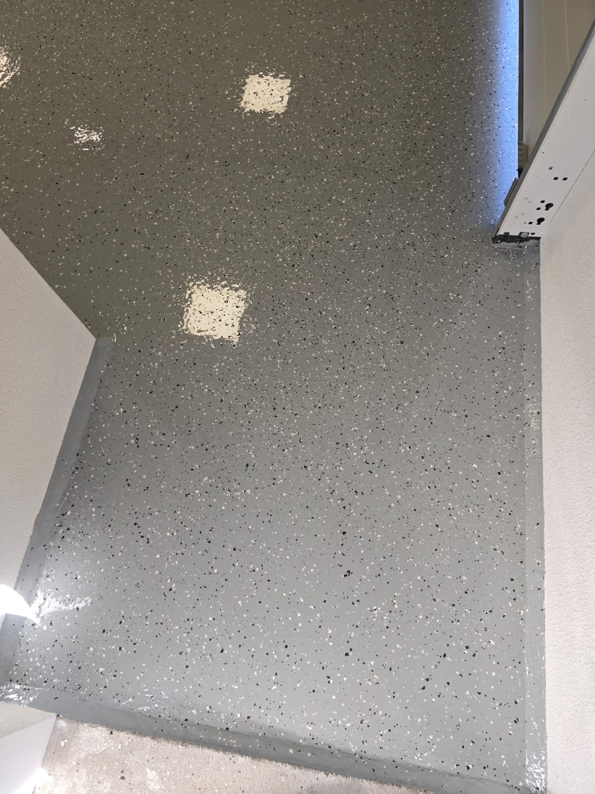 Świeżo wylana, szara posadzka z drobnymi, czarnymi i białymi plamkami w pomieszczeniu w Policach, z widocznymi odbiciami światła i narożnikiem ściany.