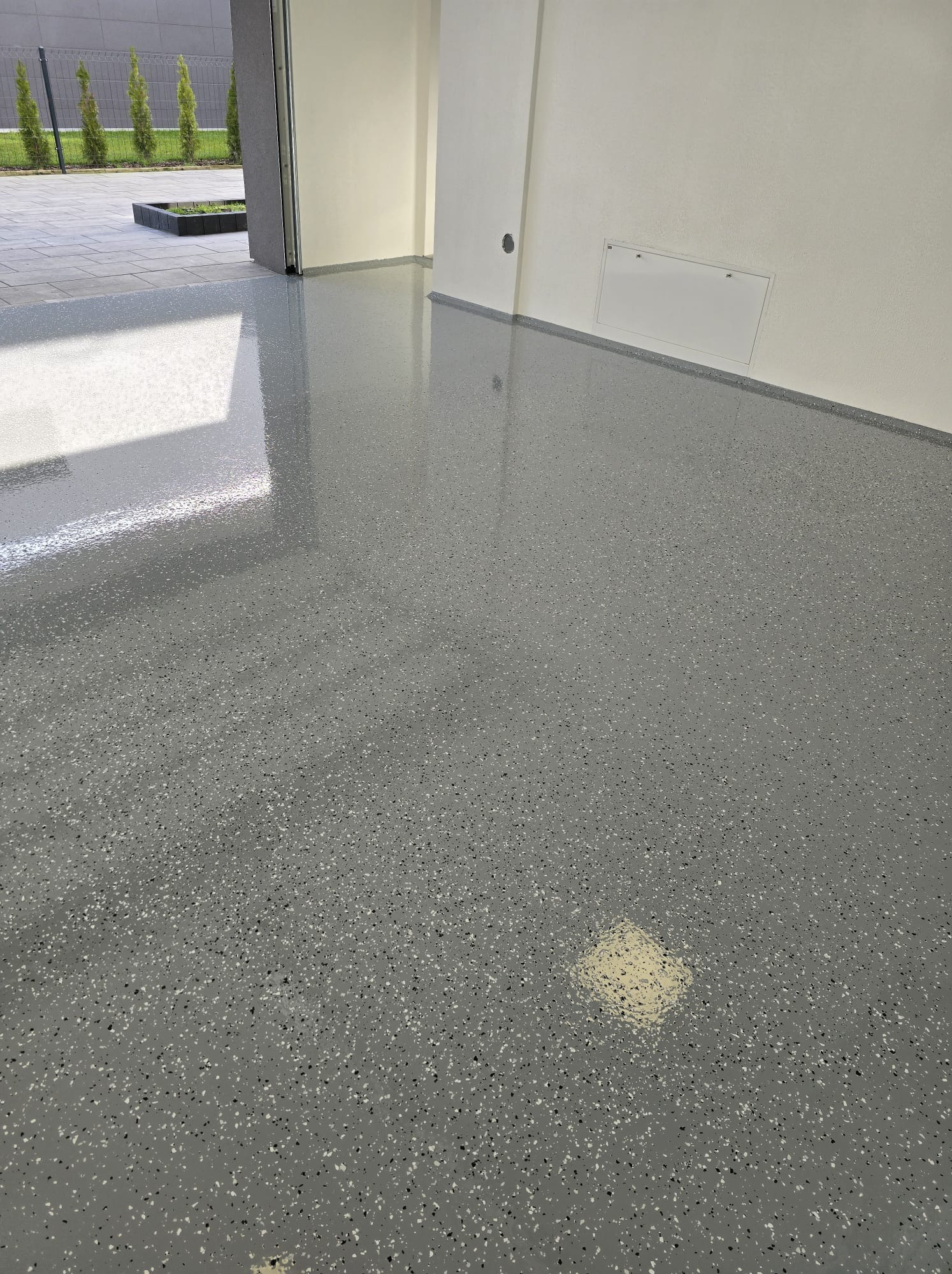 Szara, błyszcząca podłoga z drobnymi, ciemnymi i jasnymi wtrąceniami, widoczne odbicie światła z zewnątrz, przy białej ścianie z klapą rewizyjną.