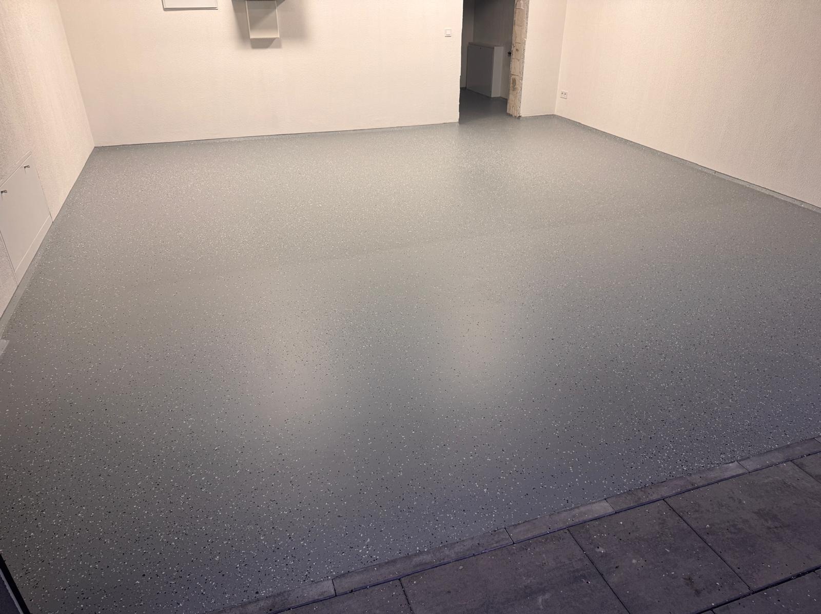 Szara podłoga w garażu pokryta żywicą z drobnymi, ciemnymi plamkami, widok na białe ściany i wejście do innego pomieszczenia.