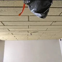 Układanie cegiełek z płyty kartonowo-gipsowej na suficie w trakcie prac wykończeniowych, widoczna instalacja elektryczna i czarna folia zabezpieczająca.