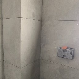 Michał Słomian firma usługowa - Świeżo ułożone, szare płytki w rogu łazienki, widoczne krzyżyki dystansowe, puszka elektryczna z przymocowaną poziomicą i wystające rury kanalizacyjne, na podłodze narzędzia.