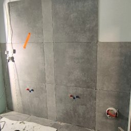 Michał Słomian firma usługowa - Szare płytki ceramiczne układane na ścianie w łazience, widoczne krzyżyki dystansowe, puszki elektryczne i rury wodne, na podłodze worek zaprawy i zielone wiadro.