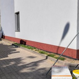 Michał Słomian firma usługowa - Ściana budynku z widoczną kamerą monitoringu, fragmentem okna i pomalowanym cokołem. Przy ścianie leżą materiały budowlane i puszki z farbą.