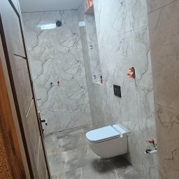 Michał Słomian firma usługowa - Nowoczesna łazienka z marmurowymi płytkami na ścianach i podłodze, wisząca toaleta z czarnym przyciskiem spłukującym, pomarańczowe zabezpieczenia na narożnikach, oświetlenie sufitowe.