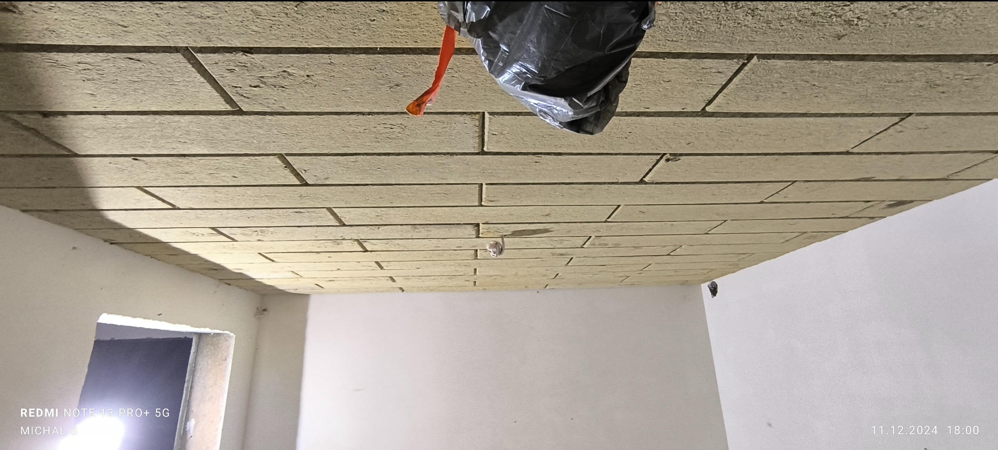 Układanie cegiełek z płyty kartonowo-gipsowej na suficie w trakcie prac wykończeniowych, widoczna instalacja elektryczna i czarna folia zabezpieczająca.