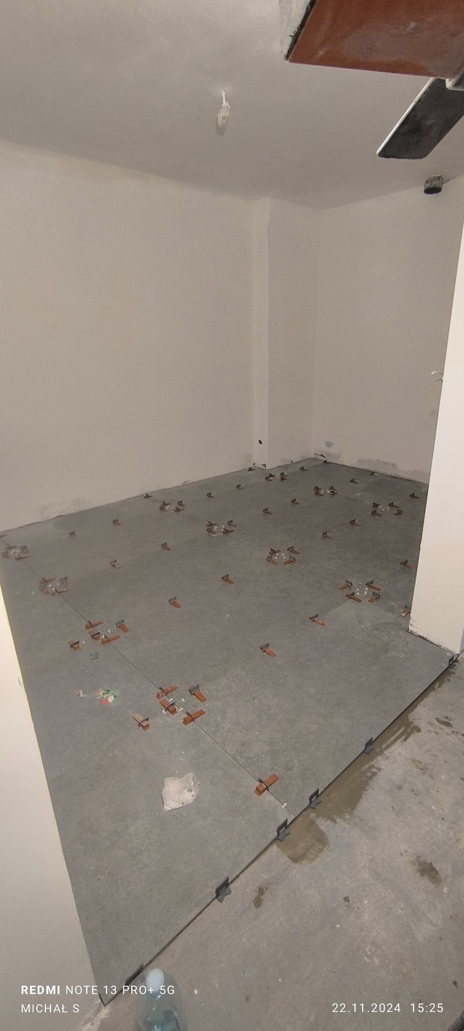 Szare płytki podłogowe w trakcie układania, z użyciem klipsów poziomujących i krzyżyków dystansowych, widok na surową ścianę i fragment sufitu z wystającym kablem.