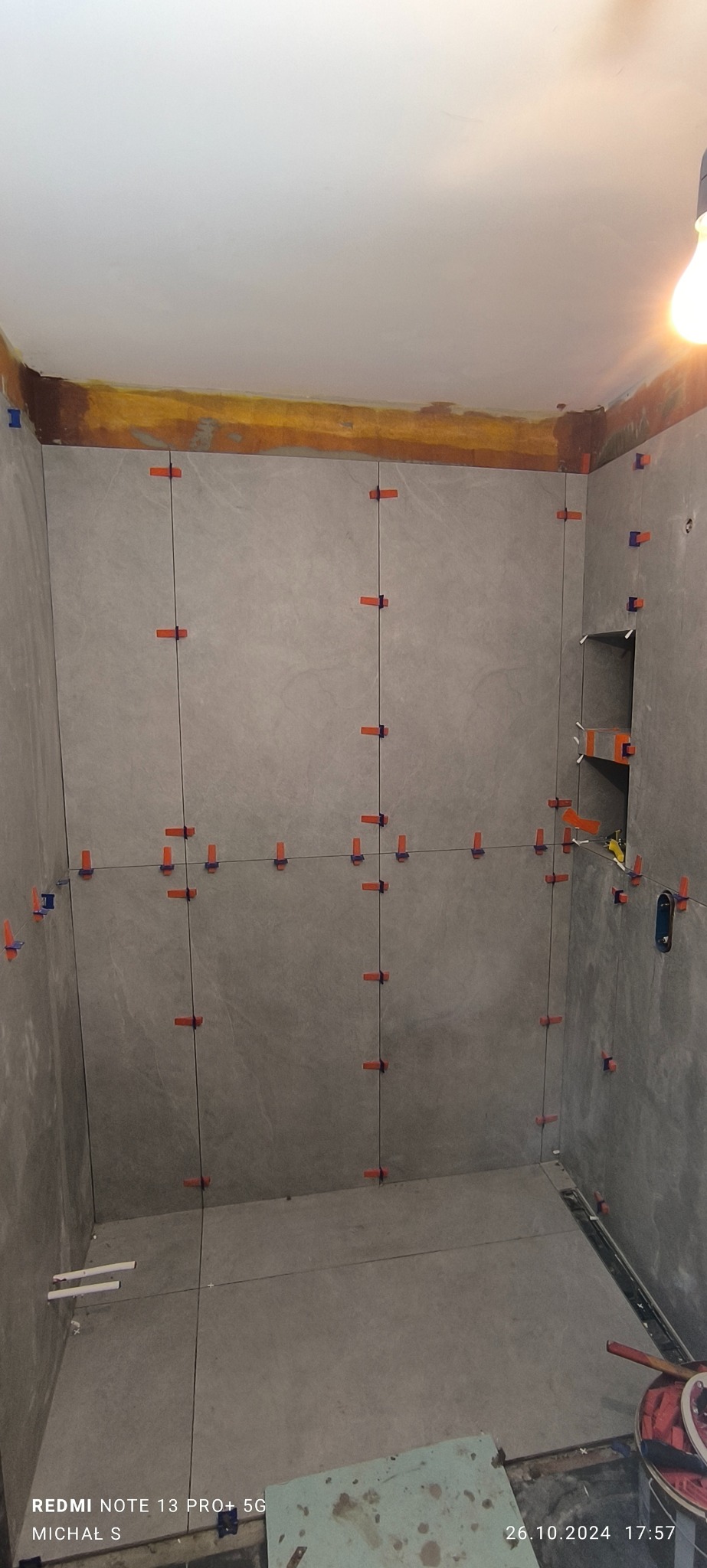 Ściany łazienki w trakcie układania szarych płytek, widoczne krzyżyki dystansowe, przygotowane półki wnękowe i fragment odpływu liniowego w podłodze.