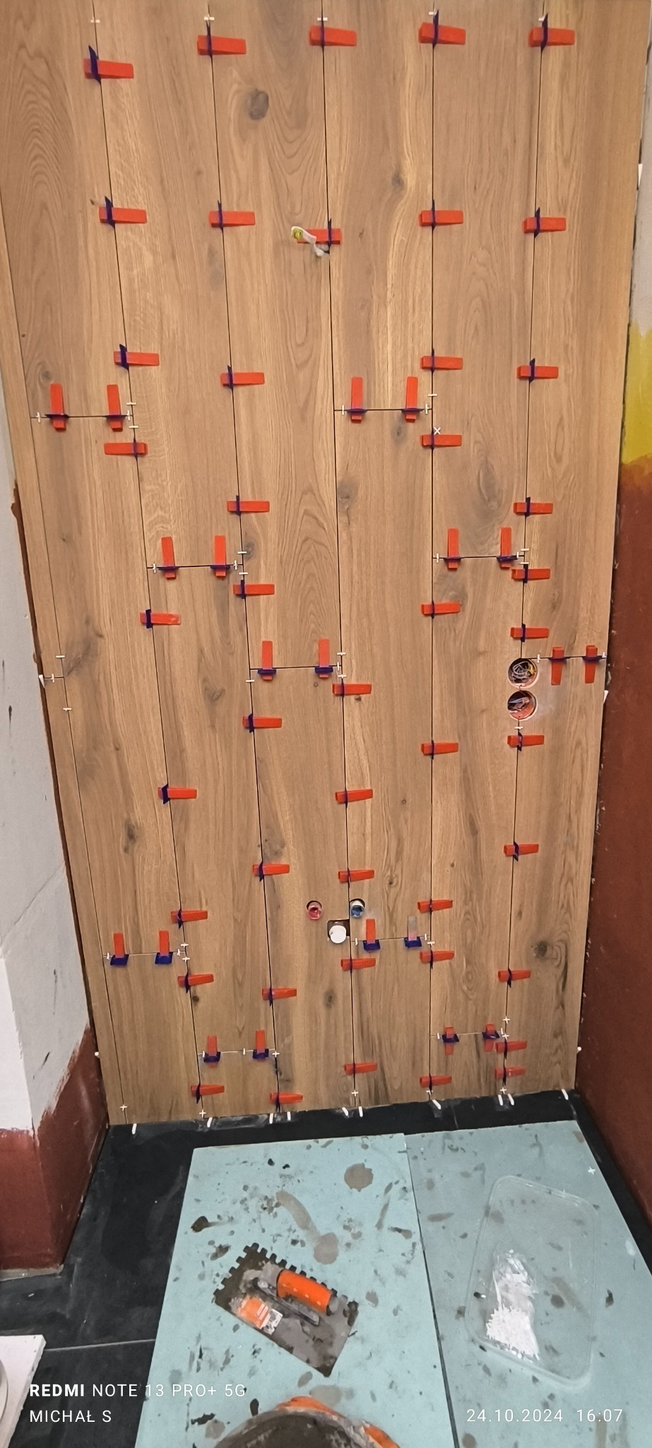 Układanie płytek imitujących drewno na ścianie z użyciem systemu poziomowania płytek, widoczne otwory na gniazdka elektryczne, na podłodze narzędzia glazurnicze.