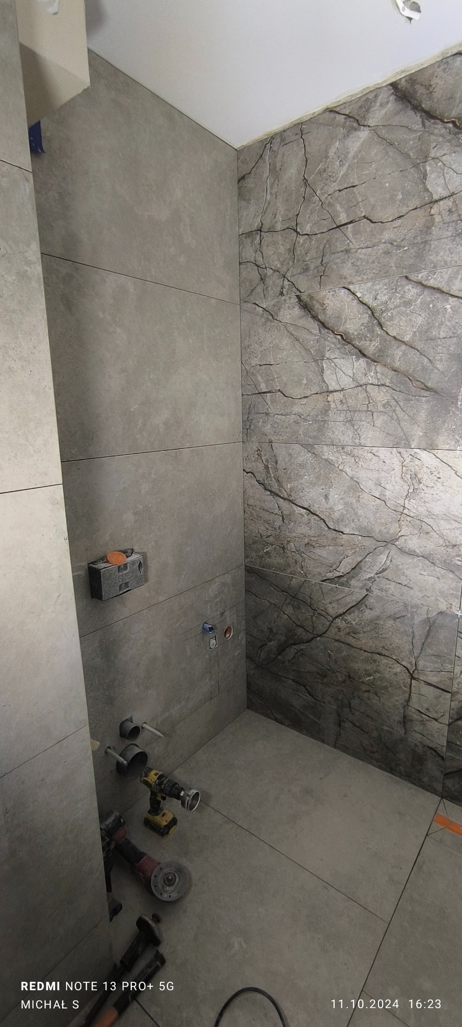 Montaż płytek imitujących marmur na ścianie w łazience, widoczne narzędzia glazurnicze i przygotowane instalacje pod armaturę.