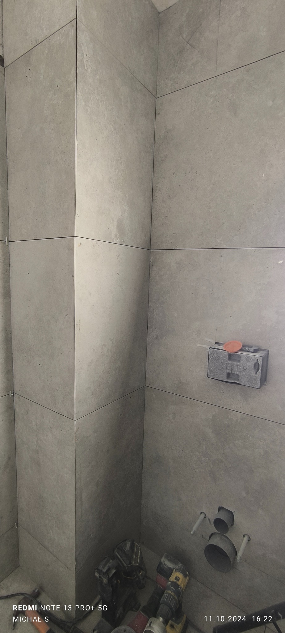 Świeżo ułożone, szare płytki w rogu łazienki, widoczne krzyżyki dystansowe, puszka elektryczna z przymocowaną poziomicą i wystające rury kanalizacyjne, na podłodze narzędzia.