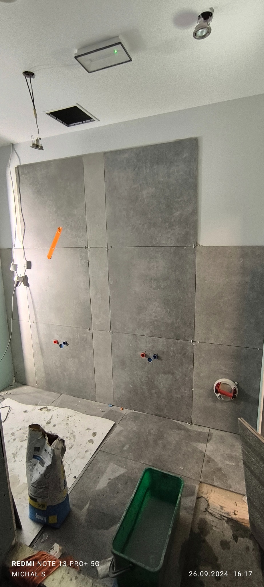 Szare płytki ceramiczne układane na ścianie w łazience, widoczne krzyżyki dystansowe, puszki elektryczne i rury wodne, na podłodze worek zaprawy i zielone wiadro.