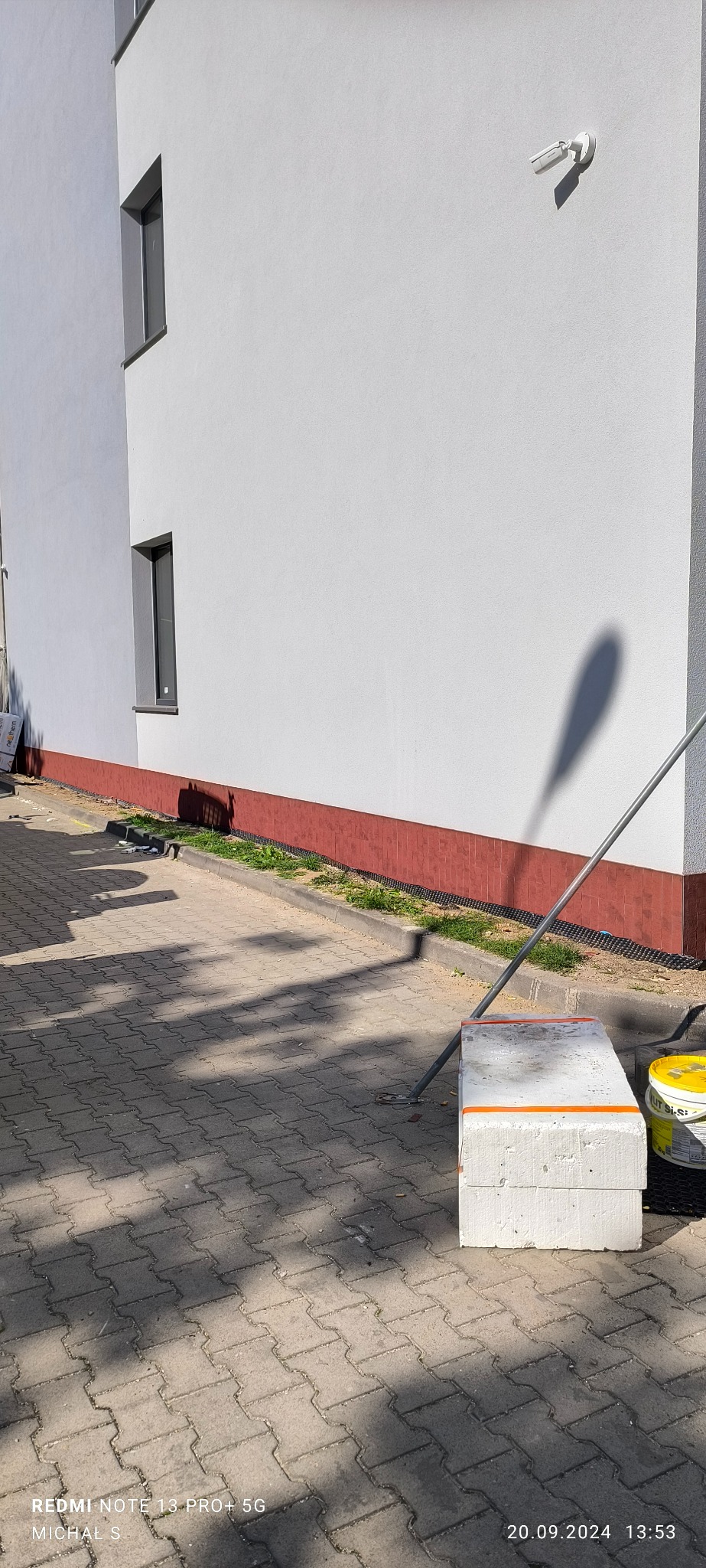 Ściana budynku z widoczną kamerą monitoringu, fragmentem okna i pomalowanym cokołem. Przy ścianie leżą materiały budowlane i puszki z farbą.