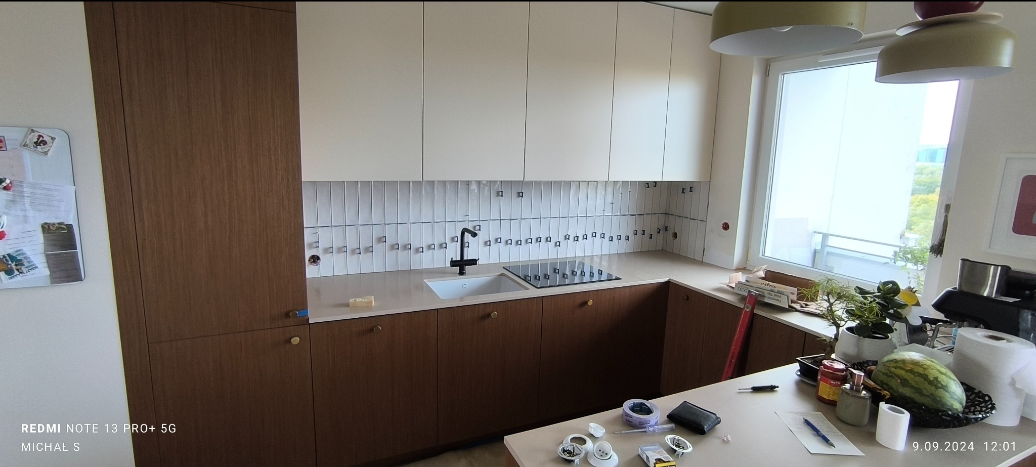 Montaż białych, wąskich płytek na ścianie nad blatem kuchennym, widoczne kleje montażowe, brązowe szafki kuchenne z okrągłymi uchwytami, czarna bateria i płyta indukcyjna, częściowo zagospodarowany...