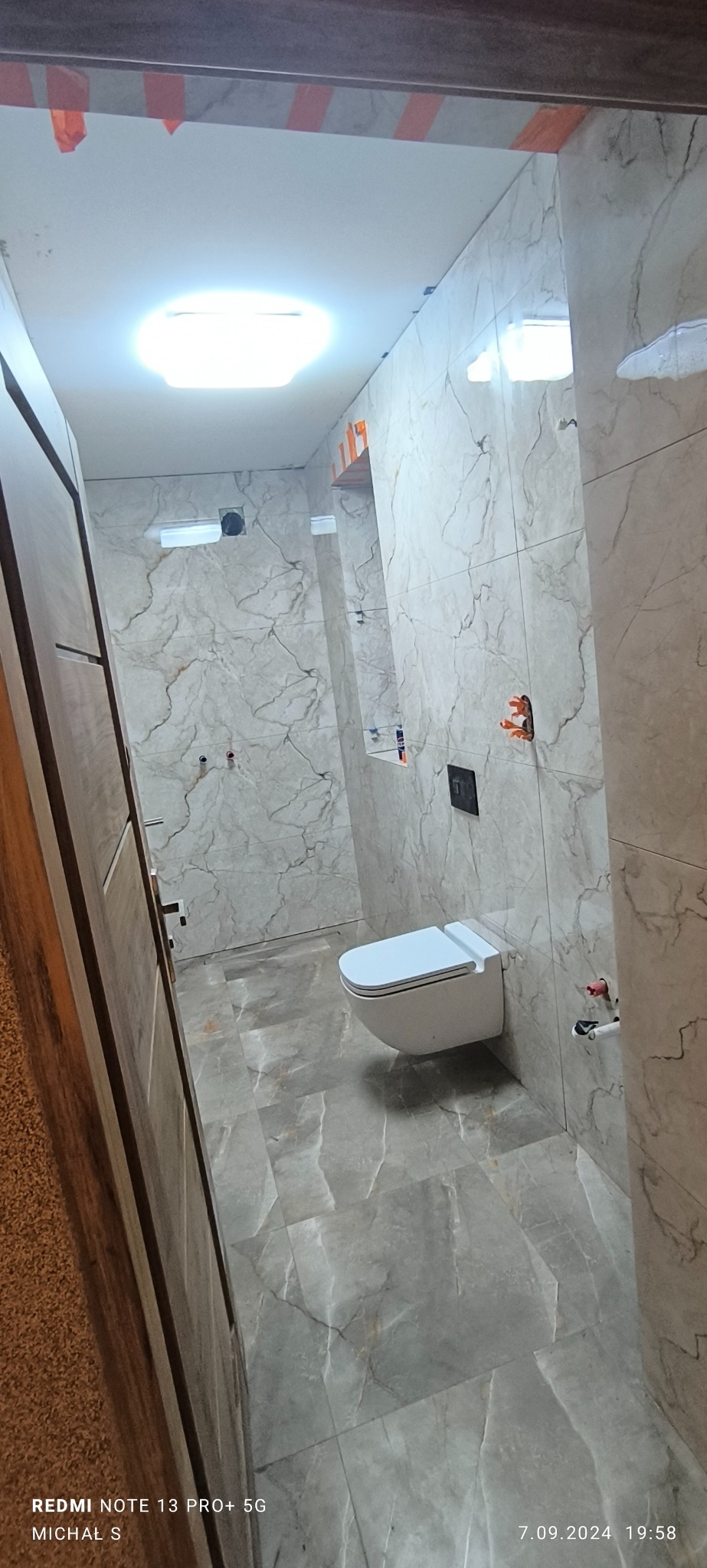 Nowoczesna łazienka z marmurowymi płytkami na ścianach i podłodze, wisząca toaleta z czarnym przyciskiem spłukującym, pomarańczowe zabezpieczenia na narożnikach, oświetlenie sufitowe.