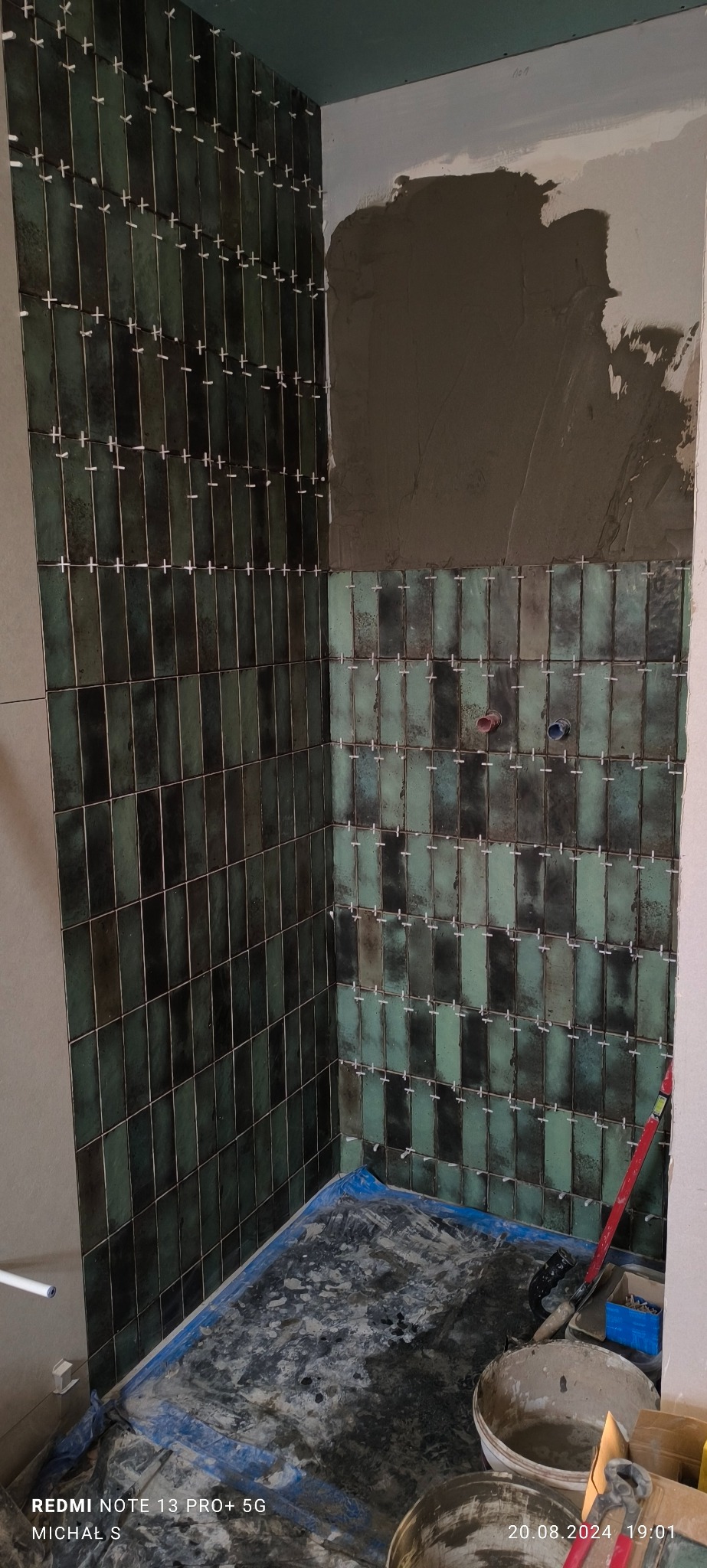 Układanie pionowych, zielono-czarnych, błyszczących płytek w łazience, widoczne krzyżyki dystansowe, fragment ściany z nałożonym klejem, zabezpieczona folią podłoga.