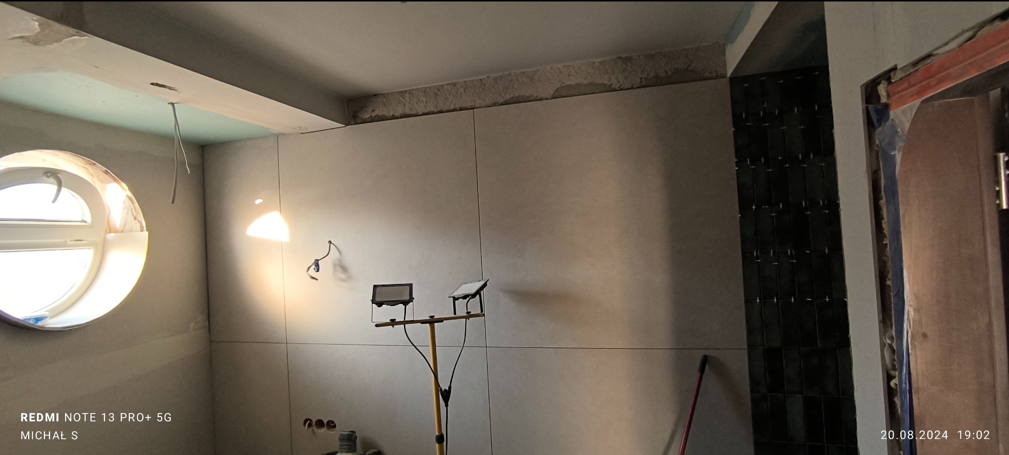 Wnętrze łazienki w trakcie prac wykończeniowych: ułożone jasne płytki wielkoformatowe, fragment ściany z ciemną mozaiką, okrągłe okno, widoczne lampy budowlane i niedokończone obramowanie drzwi.