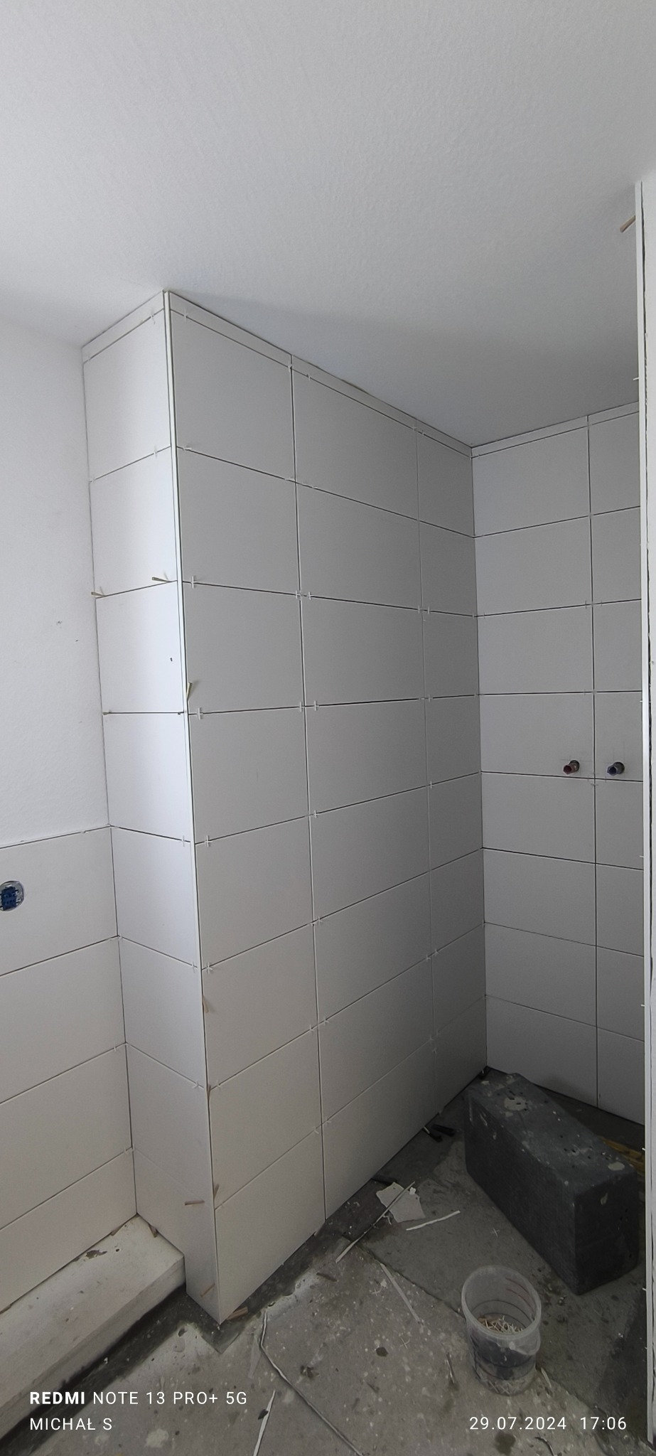 Świeżo ułożone, białe płytki na ścianach w łazience w Sierakowicach, z plastikowymi krzyżykami dystansowymi w spoinach, widoczne przygotowane instalacje wodne i resztki materiałów budowlanych...