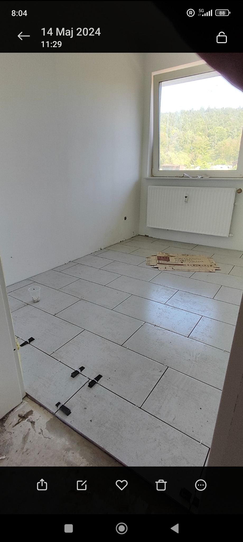 Wnętrze pomieszczenia w trakcie układania jasnych, prostokątnych płytek podłogowych; widoczne krzyżyki dystansowe, karton po płytkach i grzejnik pod oknem.