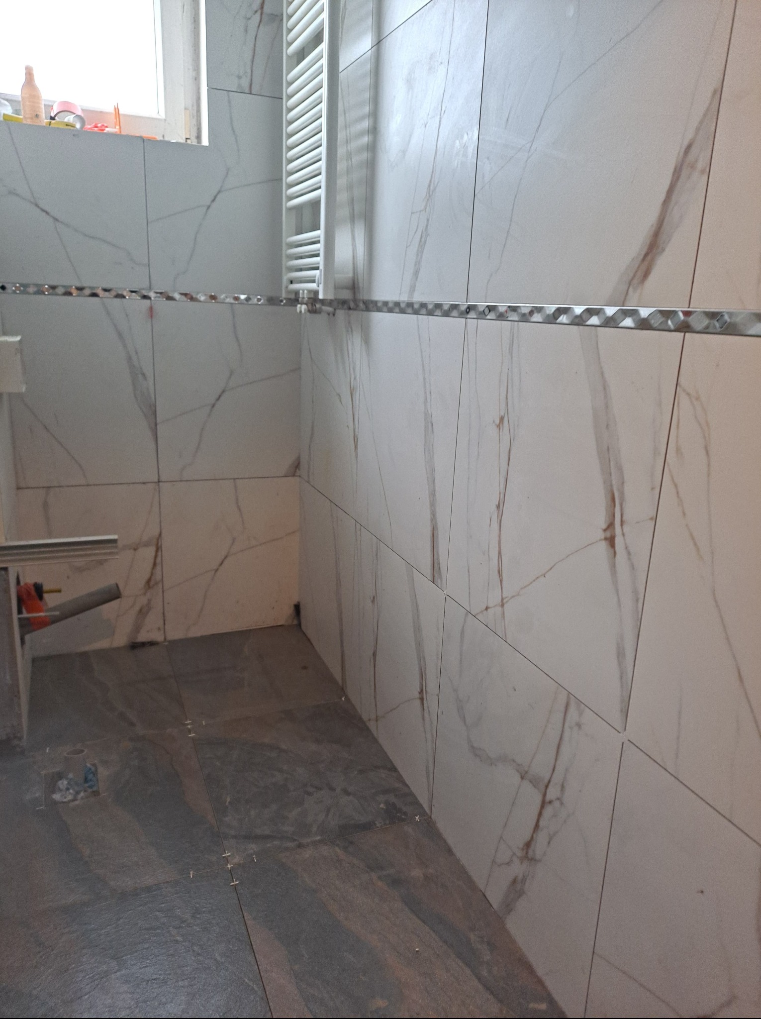 Świeżo ułożone, marmurowe płytki ścienne z dekoracyjną listwą i ciemne płytki podłogowe w trakcie remontu łazienki, widoczny grzejnik drabinkowy i narzędzia glazurnicze w rogu.