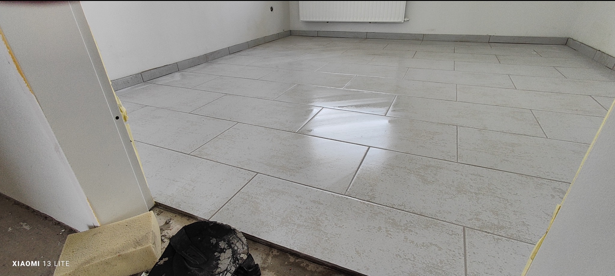 Świeżo ułożona, jasnoszara, prostokątna terakota na podłodze w pustym pomieszczeniu, widok z progu, z widocznym fragmentem pianki montażowej i czarnym workiem na gruz.
