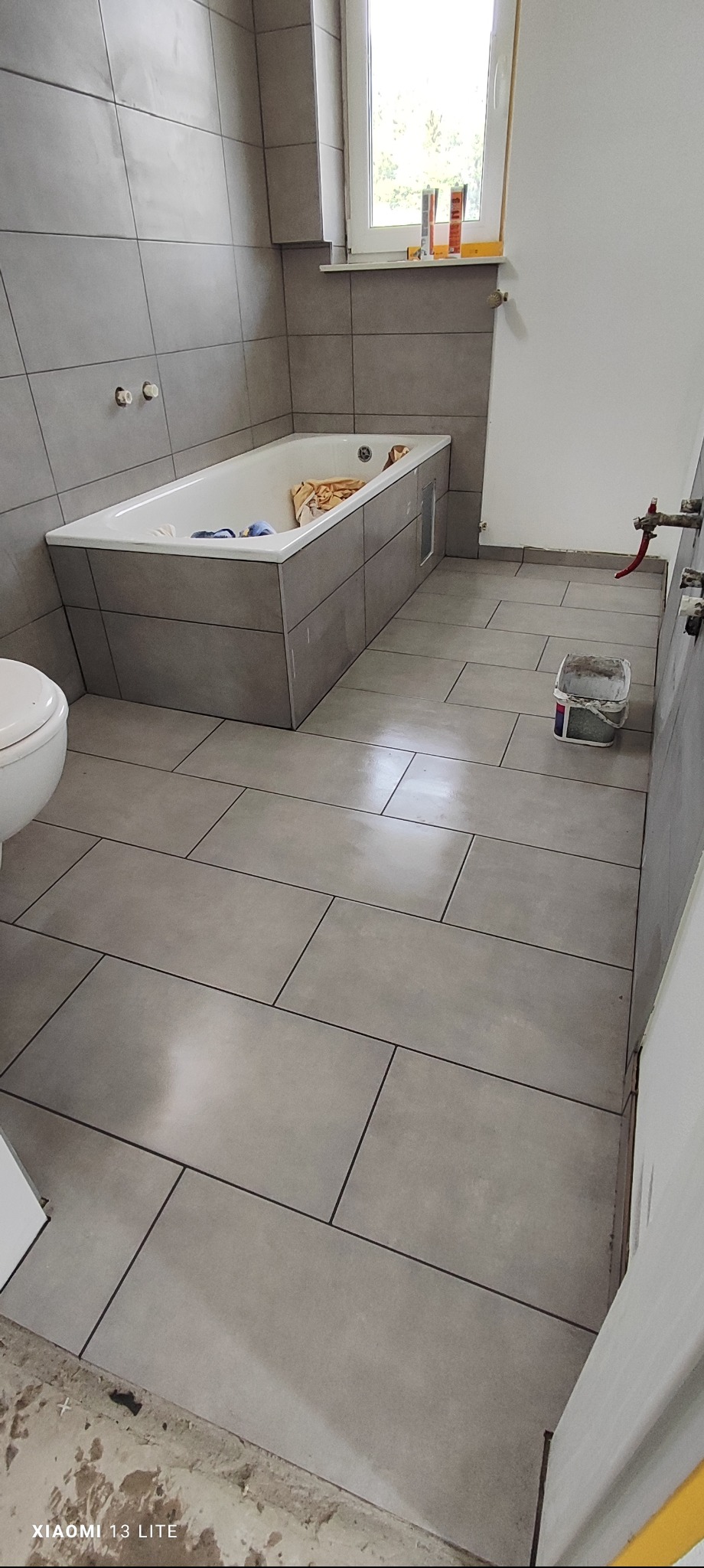 Szara łazienka w trakcie prac wykończeniowych: ułożone płytki podłogowe i ścienne, obudowana wanna, widoczne narzędzia i materiały budowlane, niedokończone detale przy drzwiach.