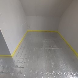 Dymitr Wasko - Przygotowanie podłoża pod ogrzewanie podłogowe: folia aluminiowa z żółtą taśmą uszczelniającą przy ścianach w pustym pomieszczeniu.