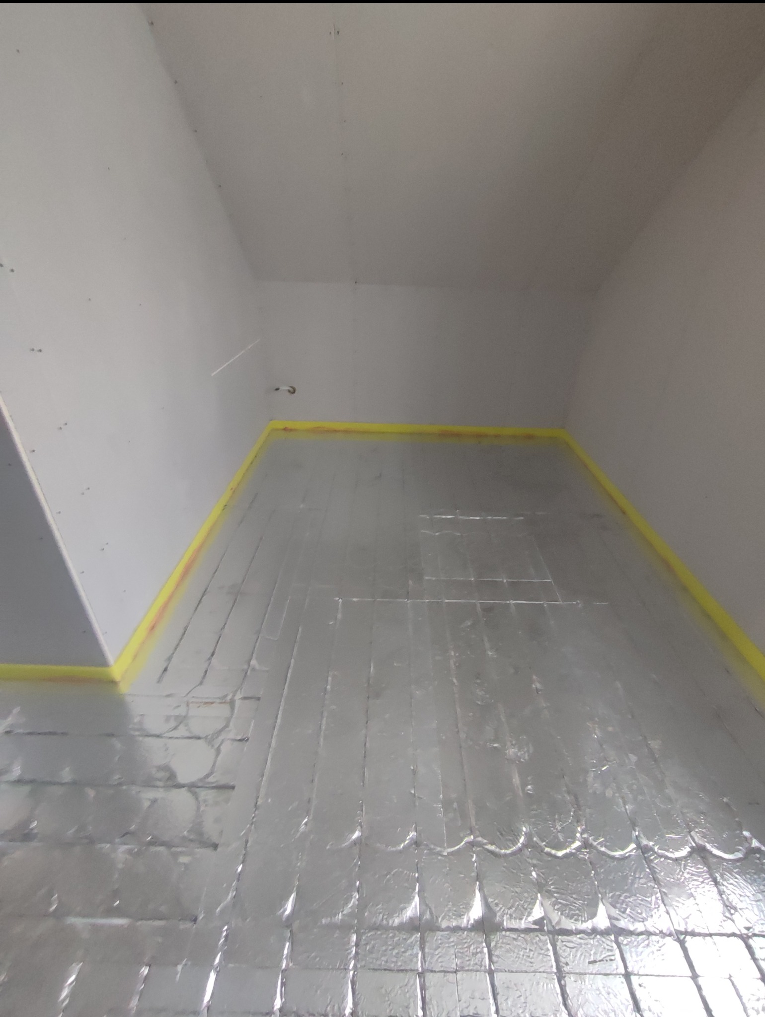 Przygotowanie podłoża pod ogrzewanie podłogowe: folia aluminiowa z żółtą taśmą uszczelniającą przy ścianach w pustym pomieszczeniu.