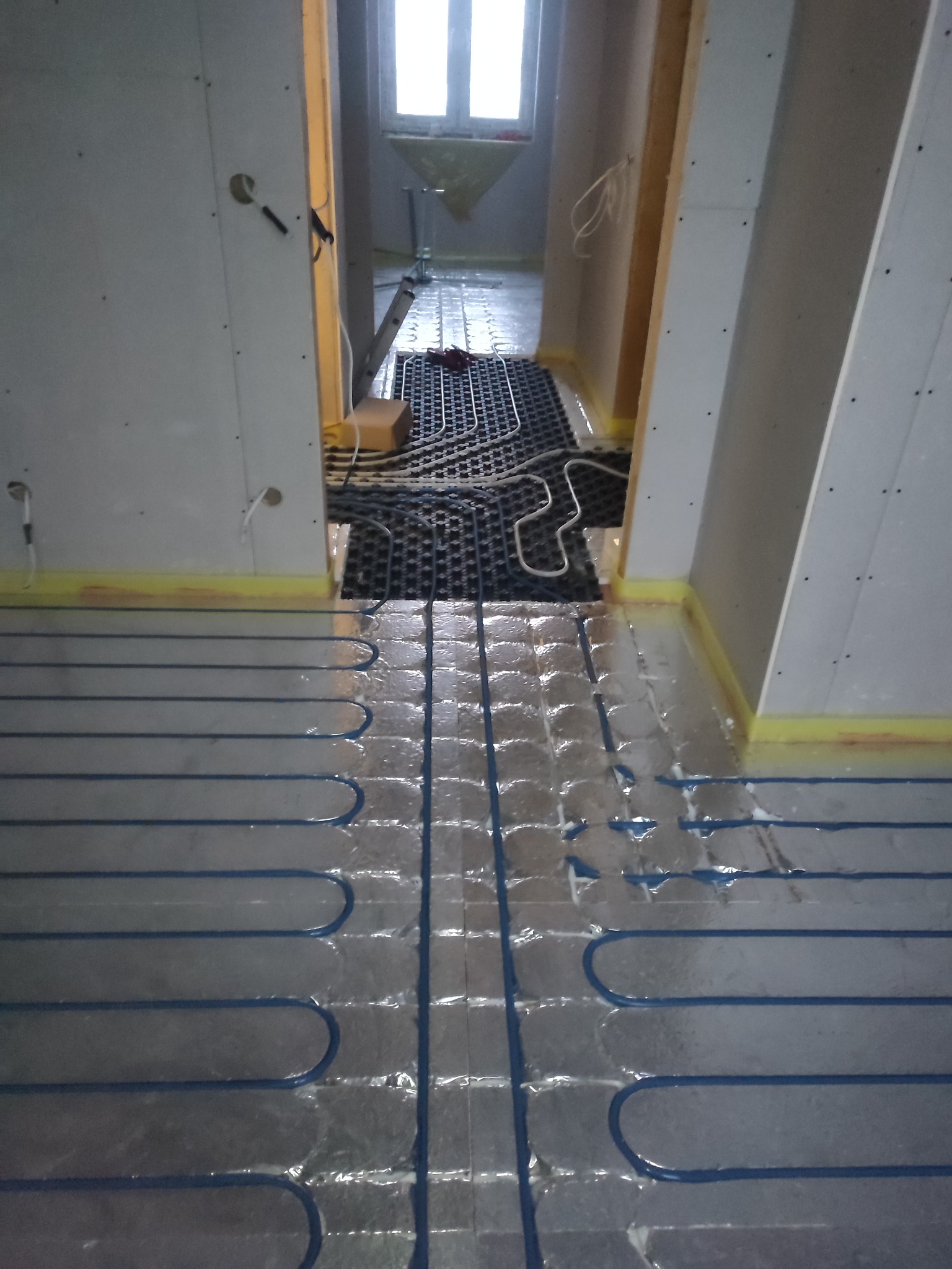 Montaż ogrzewania podłogowego: rury grzewcze w układzie meandrycznym na folii aluminiowej, widoczne fragmenty izolacji pionowej ścian, surowy stan wykończenia wnętrza.