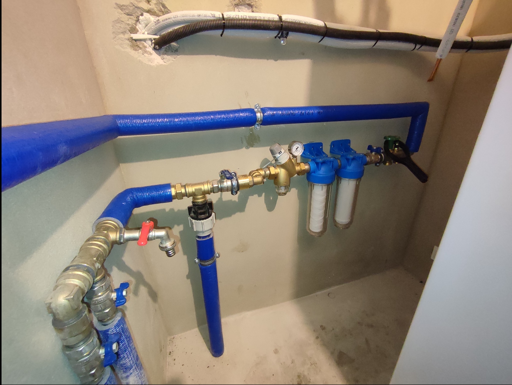 Złożona instalacja wodna z filtrami i zaworami, rury w niebieskiej izolacji termicznej, widoczne manometry i elementy metalowe, montaż w surowym pomieszczeniu z widocznymi przewodami elektrycznymi.