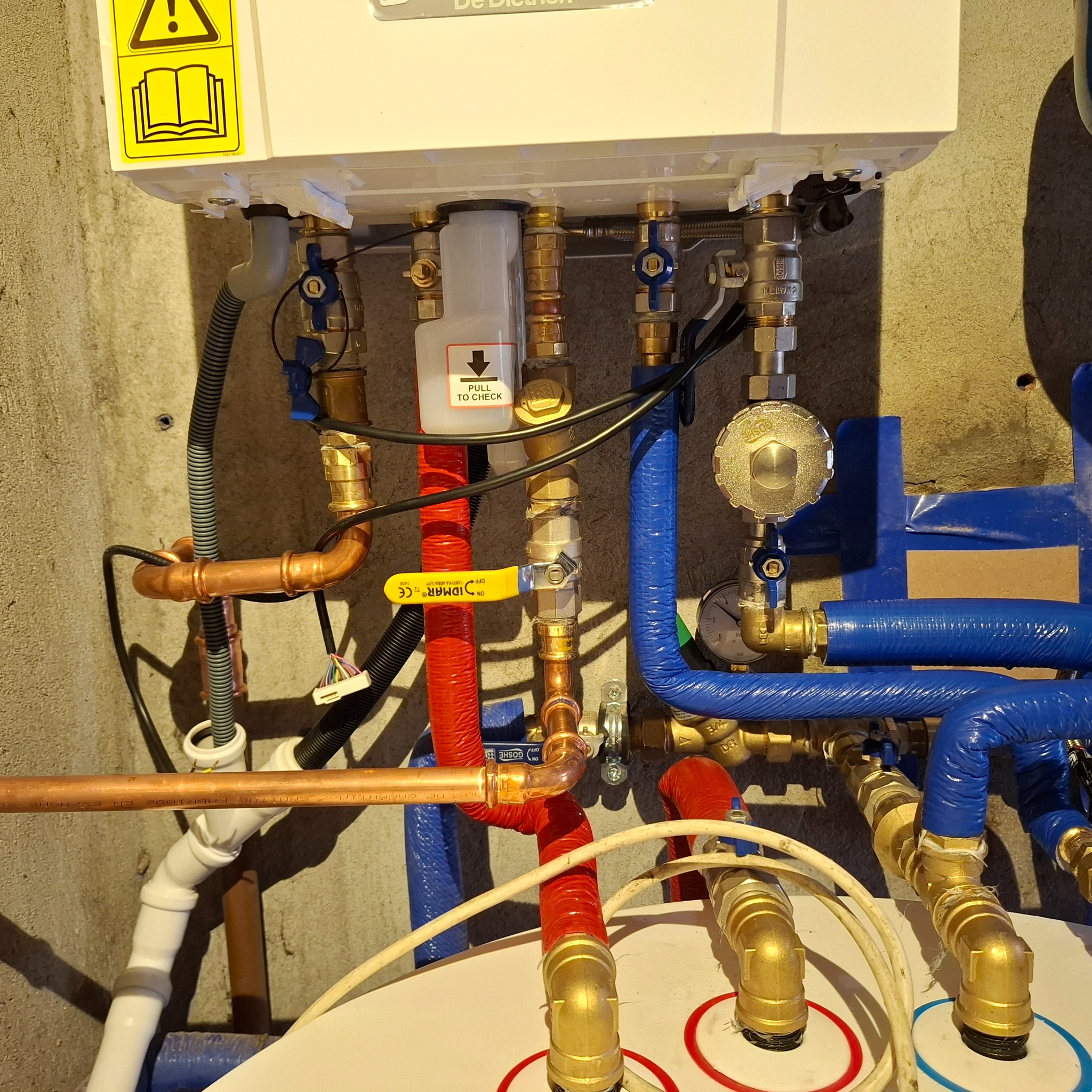 Złożona instalacja grzewcza z miedzianymi i niebieskimi rurami, zaworami i elementami pomiarowymi, zamontowana na szarej ścianie. Widoczny piec De Dietrich.