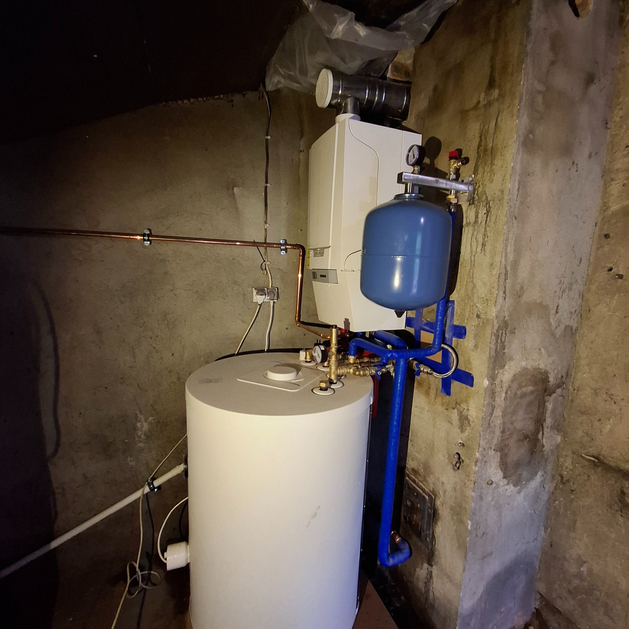 Biały piec gazowy z niebieskim zbiornikiem wyrównawczym i miedzianymi rurami w surowym pomieszczeniu. Widoczne elementy instalacji i przyłącza.