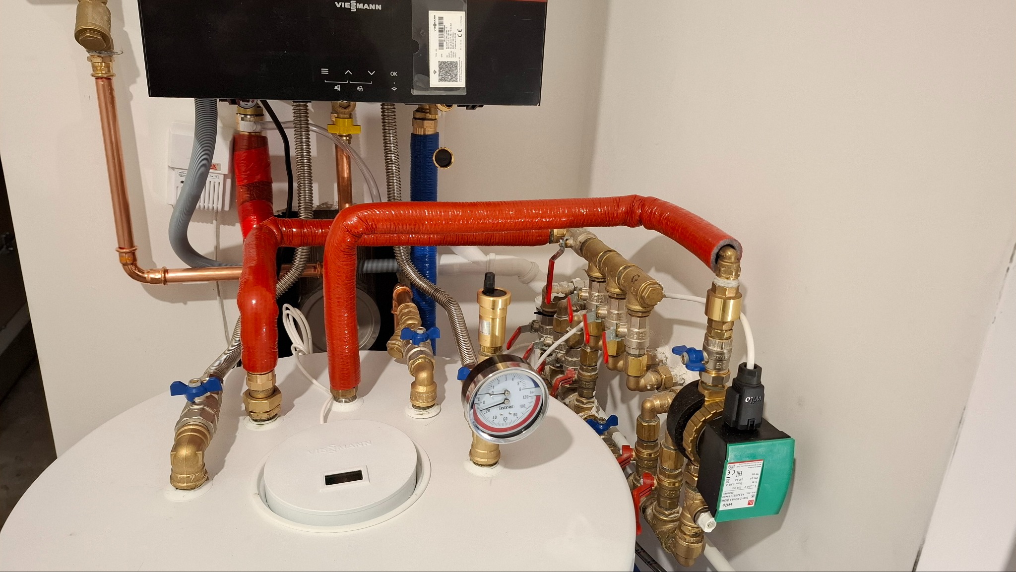 Złożona instalacja grzewcza Viessmann z czerwonymi rurami izolowanymi, manometrem i pompą obiegową Wilo, widoczna na tle białej ściany.