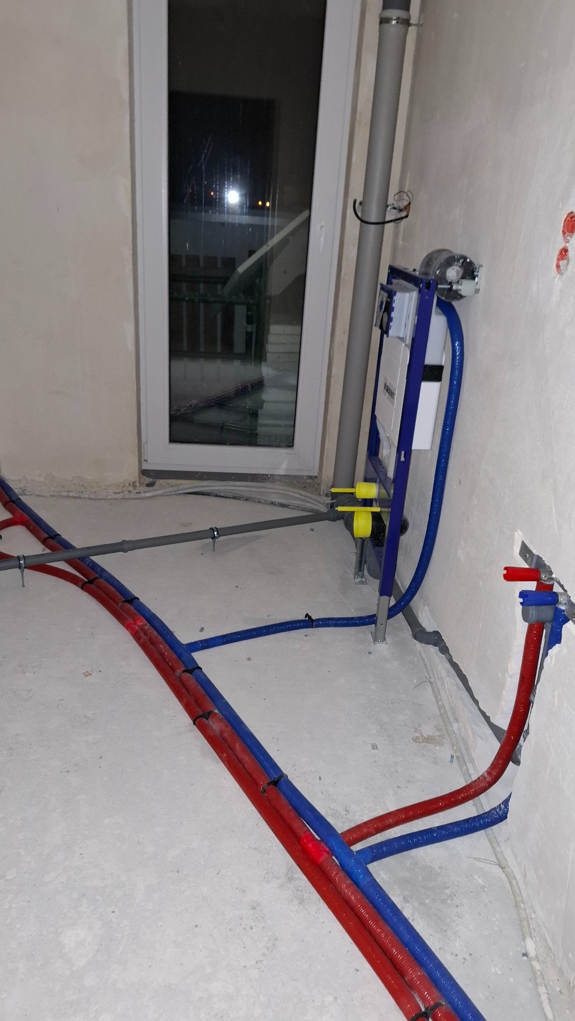 Nowa instalacja wodna z rurami w kolorze czerwonym i niebieskim, zamontowana na ścianie stelaż podtynkowy WC. Widoczne przyłącza i rury odpływowe.