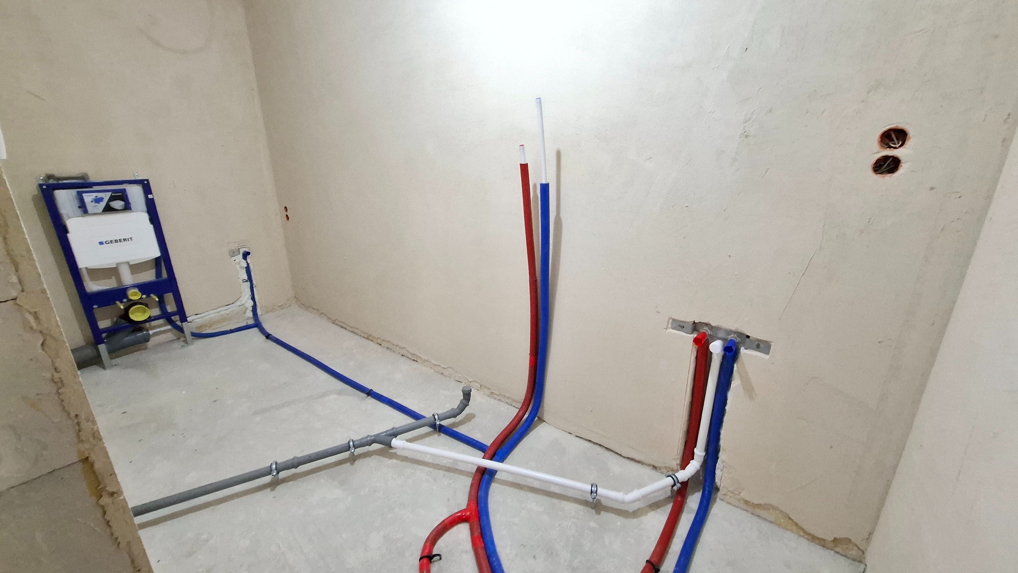 Surowy stan łazienki z instalacją wodną, widoczne rury w kolorach niebieskim i czerwonym, podłączenie do stelaża podtynkowego Geberit, ściany w trakcie wykończenia.