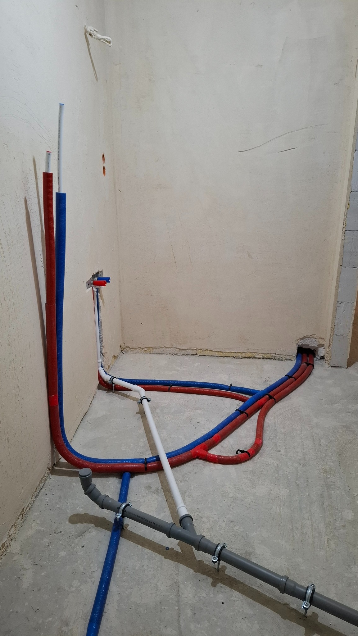 Instalacja wodna w trakcie montażu: rury PEX w kolorach niebieskim i czerwonym ułożone na surowej posadzce, przyłącza do ściany. Widoczne elementy kanalizacji.