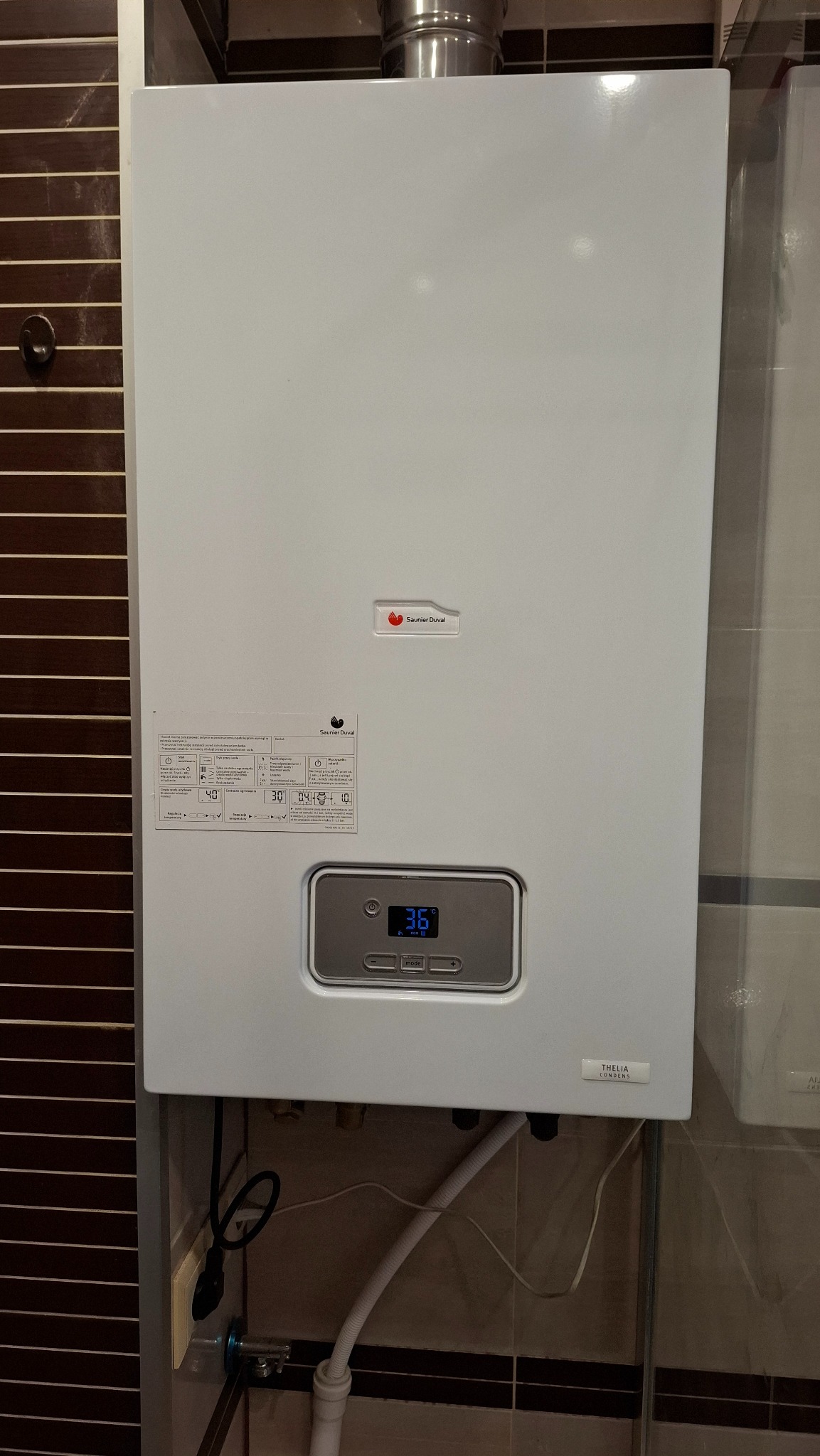 Biały, wiszący piec gazowy Saunier Duval Thelia Condens, z wyświetlaczem wskazującym 36 stopni, zamontowany na ścianie z brązowo-białymi płytkami.