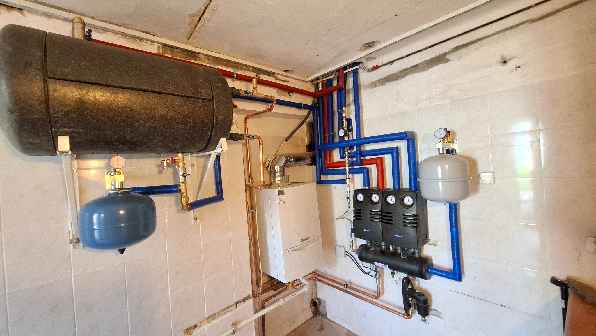 Kompleksowa instalacja grzewcza z kotłem Vaillant, zasobnikiem ciepłej wody i grupą pompową na tle białych płytek. Rury miedziane i izolowane na ścianie.