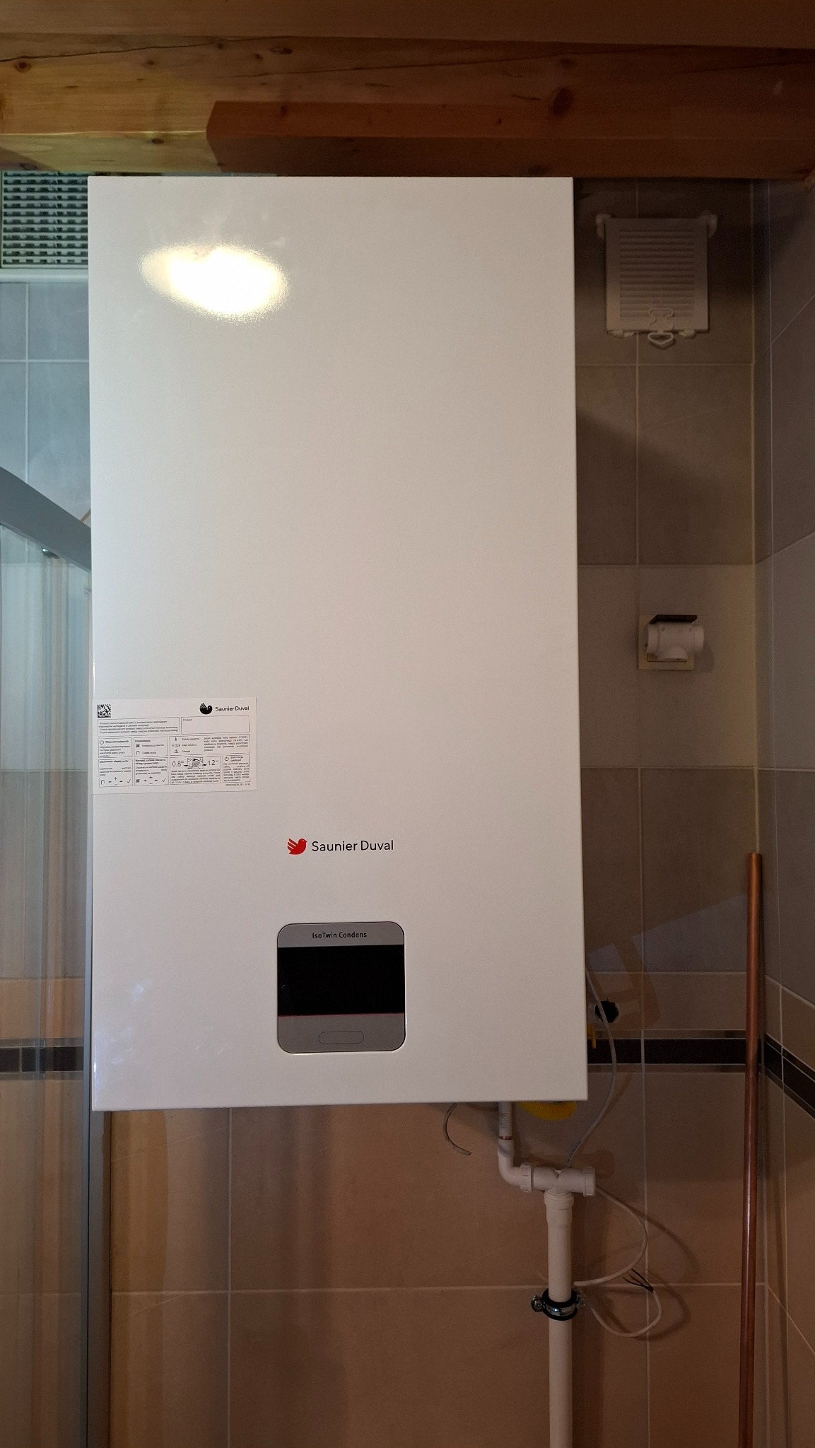 Biały, wiszący piec gazowy Saunier Duval IsoTwin Condens na tle beżowych płytek. Widoczne rury instalacyjne i element drewnianej konstrukcji stropu.